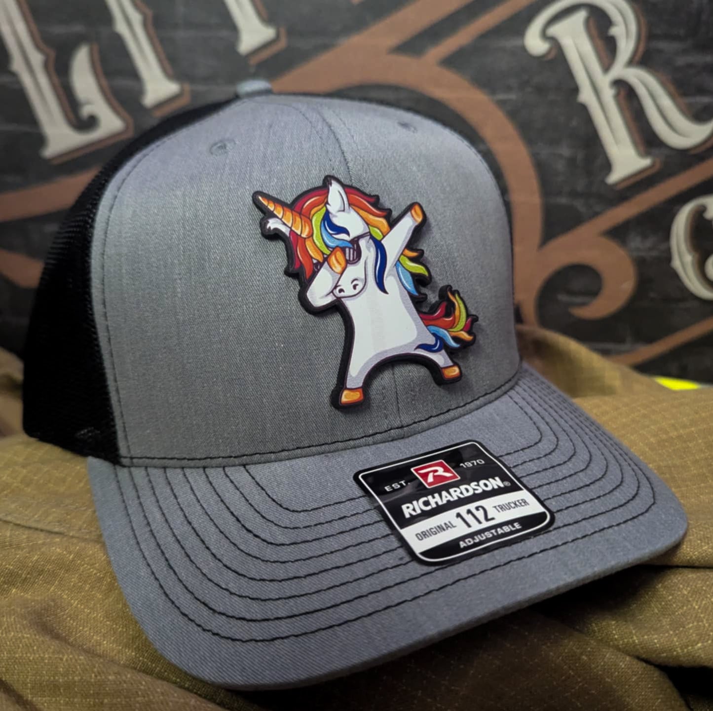 Richardson hat with leather patch of rainbow unicorn doing a dab pose on gray and black trucker hat