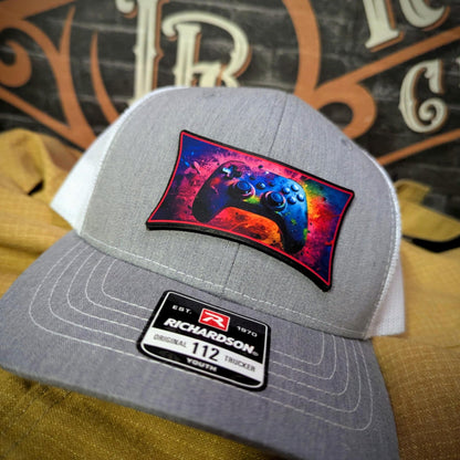 Richardson hat with full-color leather patch of video game controller in neon splatter design on gray and white trucker hat